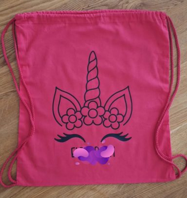 nachhaltige Geschenkverpackung Roter Rucksack mit einem Einhorn-Motiv und blühenden Augen und einem bunten Herzen.