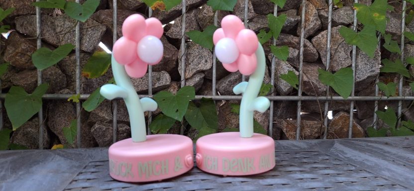 LED-Lampe mit Vinyl-Aufschrift Zwei stilisierte Blumen mit rosa Blüten und grünen Stängeln auf einem pinken Sockel.